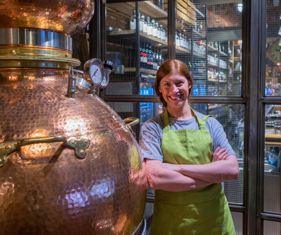 Woman who is smiling leans against copper distilling machine